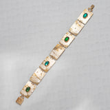 The Gilded Petra Emerald Bracelet by Holly Yashi showcases rectangular textured links adorned with oval emerald green crystals. The handcrafted bracelet is shown diagonally on a light gray patterned surface.