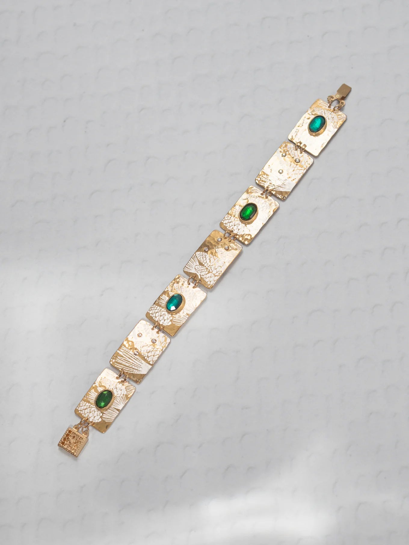 The Gilded Petra Emerald Bracelet by Holly Yashi showcases rectangular textured links adorned with oval emerald green crystals. The handcrafted bracelet is shown diagonally on a light gray patterned surface.