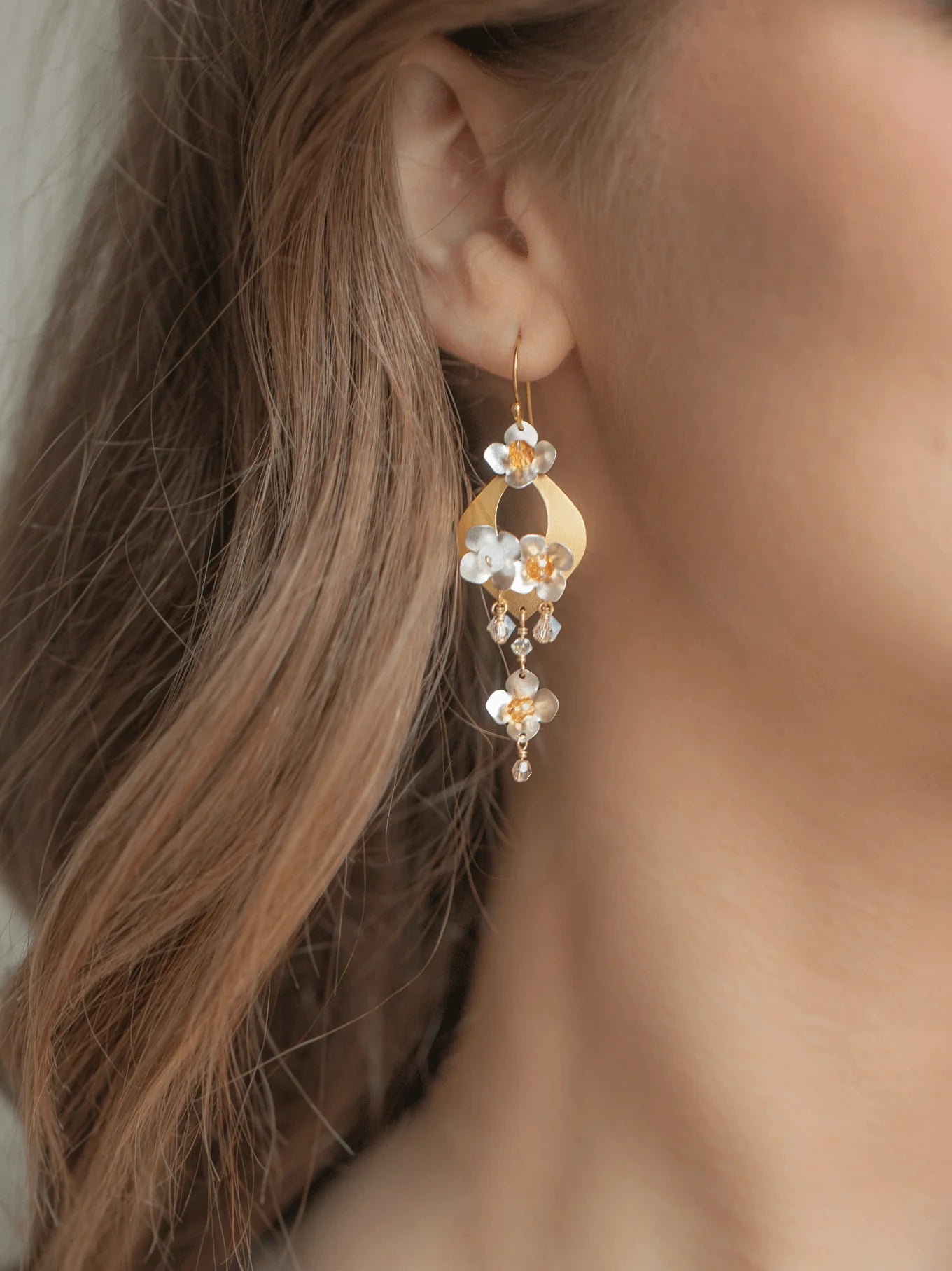 Close-up of a woman's ear wearing Holly Yashi's Hawaiian Wedding Gold/Silver Earrings—dangle earrings with white and amber flower-shaped beads. Her wavy brown hair beautifully frames the elegant Holly Yashi jewelry.