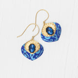 The Arabella Sapphire Blue Earrings by Holly Yashi feature gold hooks, blue and gold accents, a central shiny blue gem, and a patterned blue background displayed on a white wood surface.