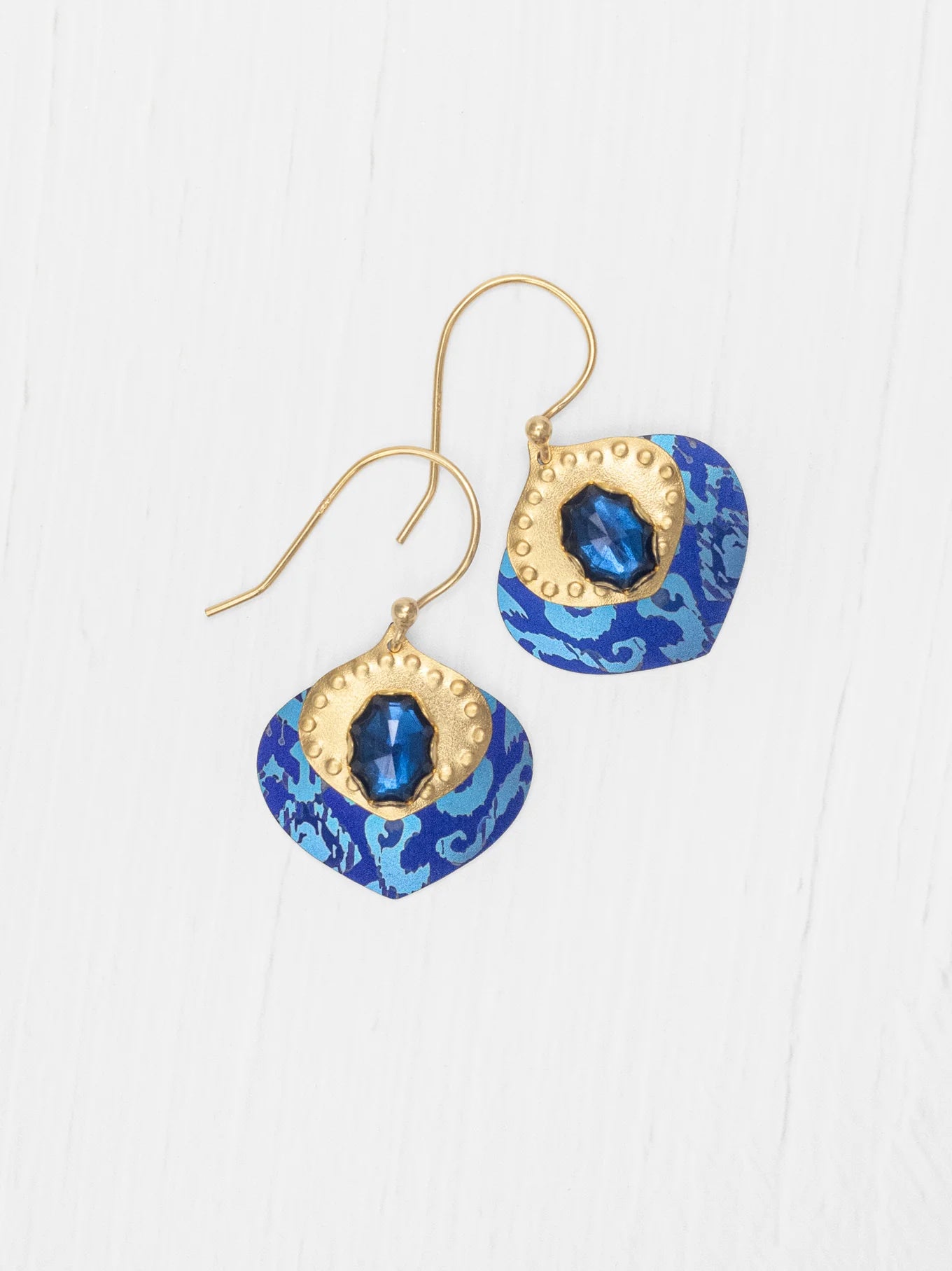 The Arabella Sapphire Blue Earrings by Holly Yashi feature gold hooks, blue and gold accents, a central shiny blue gem, and a patterned blue background displayed on a white wood surface.