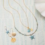Two gold necklaces with star and seashell pendants are laid on a textured surface. Alongside are Holly Yashis Carmel Seashore Blue Starfish Earrings, featuring hypo-allergenic, colorful niobium with star and shell designs. The jewelry is in blue and gold hues, with a seashell nearby.