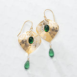 The Estelle Emerald Earrings by Holly Yashi feature hammered gold ovals with a central green gemstone and a dangling green teardrop, shown on a light textured surface.