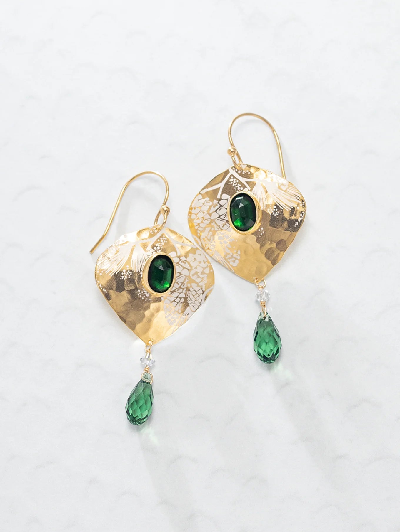 The Estelle Emerald Earrings by Holly Yashi feature hammered gold ovals with a central green gemstone and a dangling green teardrop, shown on a light textured surface.