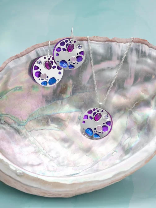 Displayed on a large open seashell against a light turquoise background, the Ocean Depth Sea Plum Earrings by Holly Yashi feature circular pendants with purple and blue Austrian crystals. These hypoallergenic earrings combine style and comfort.
