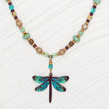 Introducing the Turquoise Beaded Dragonfly Dreams Necklace by Holly Yashi: a stunning piece with earth-toned beads and a vibrant dragonfly pendant. The niobium wings shimmer in deep blue and turquoise on a textured white background, creating an eye-catching, hypo-allergenic accessory.