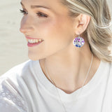 A smiling woman with light hair wears matching circular pendant earrings and the stunning Ocean Depths Sea Plum Necklace by Holly Yashi, both featuring purple and blue Austrian crystals. She elegantly pairs them with a white top, glancing slightly to the side against a soft background.