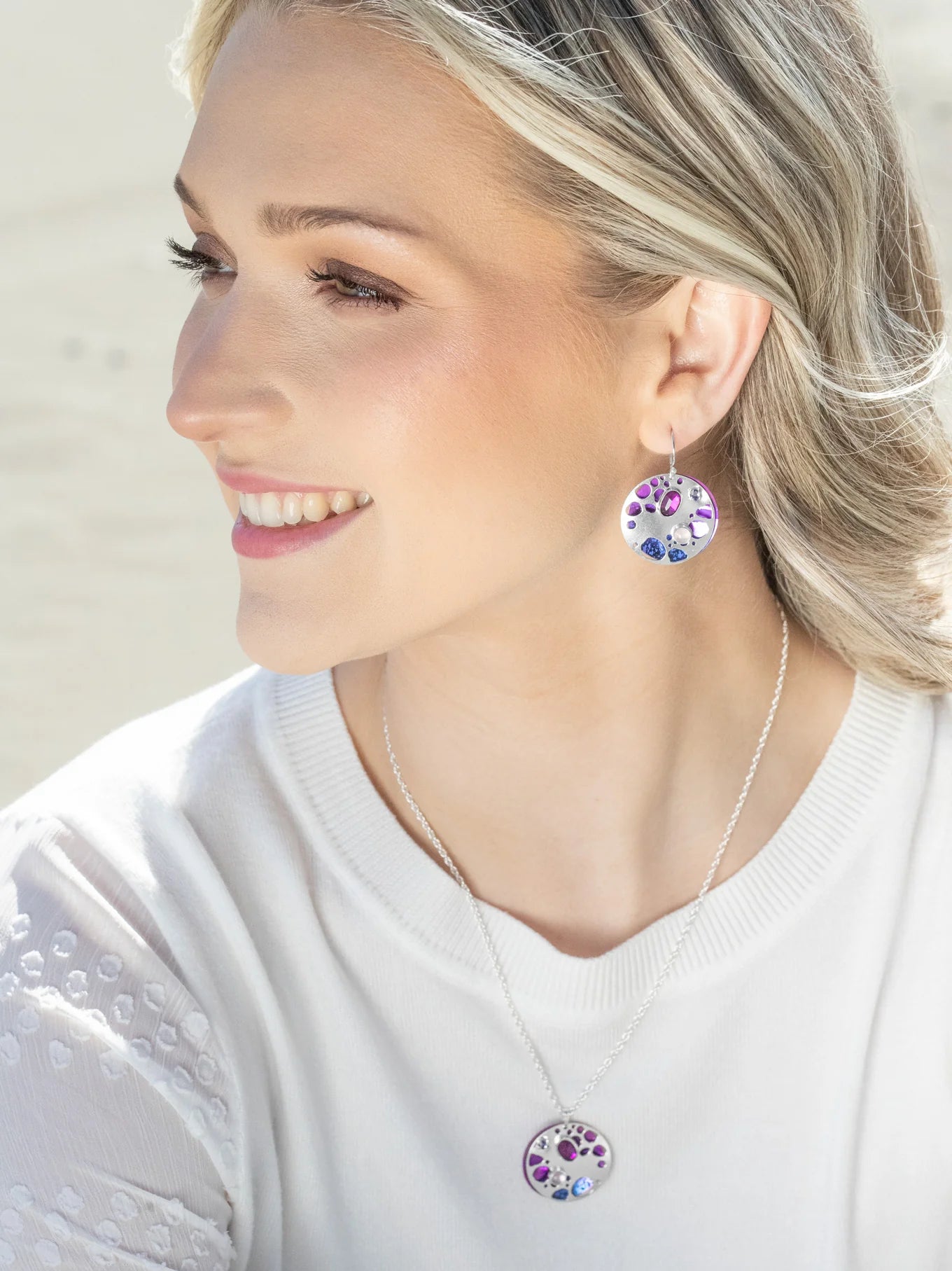 A smiling woman with light hair wears matching circular pendant earrings and the stunning Ocean Depths Sea Plum Necklace by Holly Yashi, both featuring purple and blue Austrian crystals. She elegantly pairs them with a white top, glancing slightly to the side against a soft background.