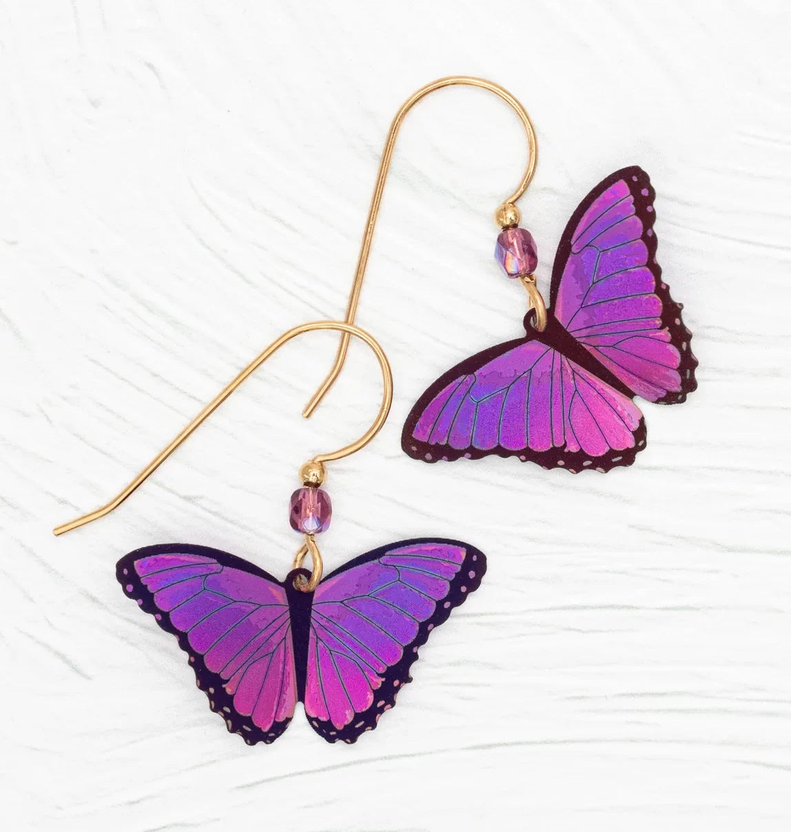 The Ultra Violet Bella Butterfly Earrings by Holly Yashi showcase two handcrafted earrings with butterfly charms in pink-to-purple hues, each on a gold hook with a purple bead, all set against a textured white background.