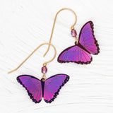 The Ultra Violet Bella Butterfly Earrings by Holly Yashi showcase two handcrafted earrings with butterfly charms in pink-to-purple hues, each on a gold hook with a purple bead, all set against a textured white background.