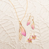 The Holly Yashi Special Edition Flutterby Pink Lariat Necklace and two pairs of matching earrings—one with pink and gold wing motifs, the other with pink flowers—are displayed on a textured, light surface.