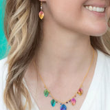 A woman smiles brightly, wearing a gold necklace and Healing Leaf Peach Earrings by Holly Yashi, with multicolored leaf pendants. Her wavy hair falls over a white top against a vibrant turquoise backdrop. The hypo-allergenic niobium ensures both comfort and style.