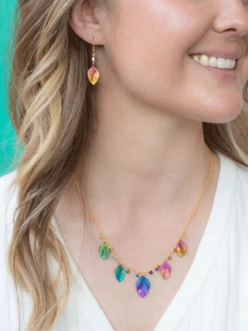 A woman smiles brightly, wearing a gold necklace and Healing Leaf Peach Earrings by Holly Yashi, with multicolored leaf pendants. Her wavy hair falls over a white top against a vibrant turquoise backdrop. The hypo-allergenic niobium ensures both comfort and style.