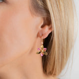 A close-up of a womans ear wearing the Special Edition Orla Drops Earrings by Holly Yashi, featuring dangling flower-shaped designs with pink Austrian crystal petals and green leaves. She has straight blonde hair and light skin.