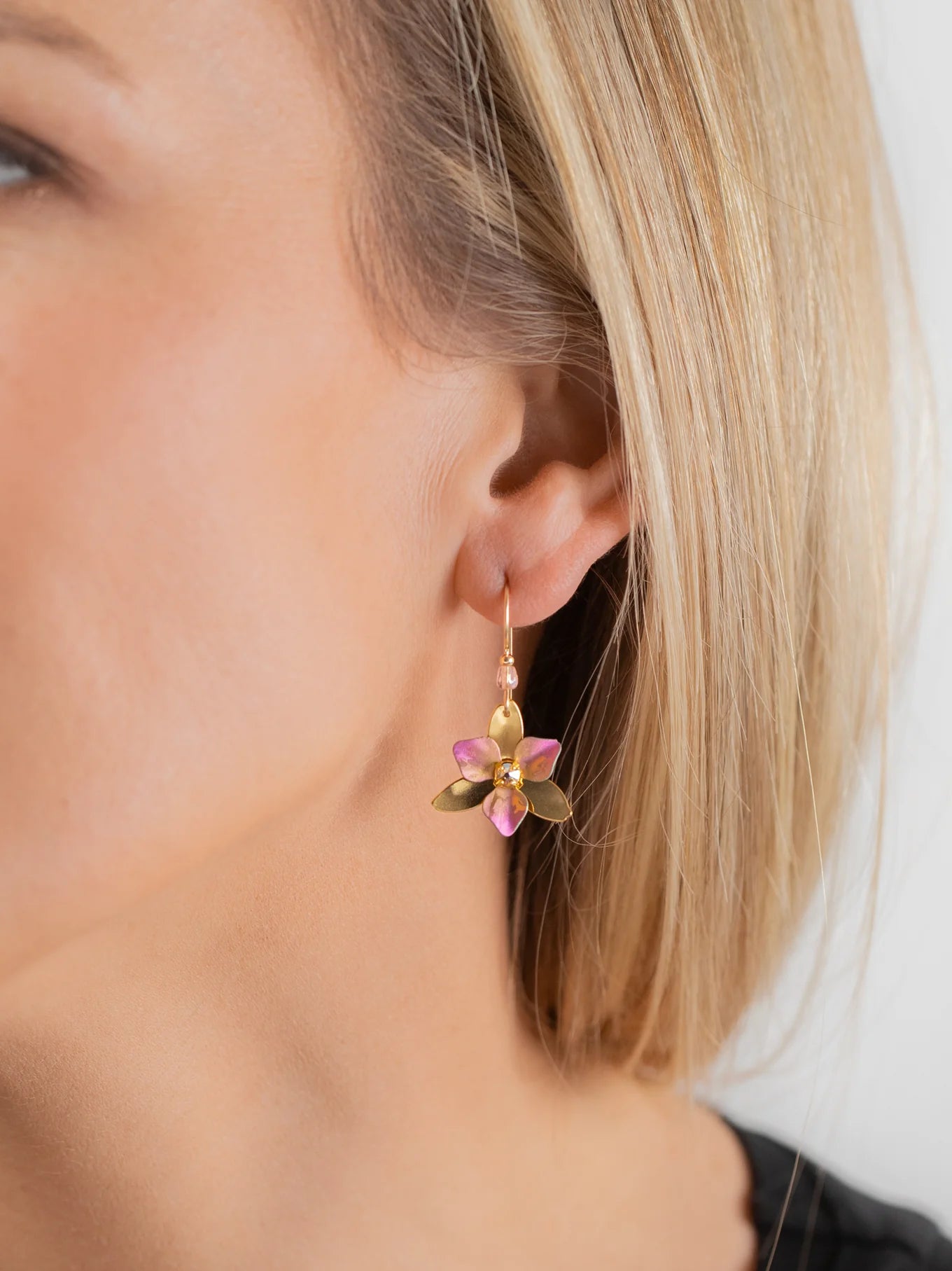 A close-up of a womans ear wearing the Special Edition Orla Drops Earrings by Holly Yashi, featuring dangling flower-shaped designs with pink Austrian crystal petals and green leaves. She has straight blonde hair and light skin.