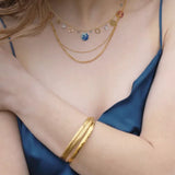 A individual in a silky blue sleeveless dress adorns layered Coral Reef Necklaces by Holly Yashi, featuring gold-plated chains with small charms. A gold bangle enhances their wrist, exuding oceanic elegance. The image is cropped to highlight the grace of the upper body and arms.