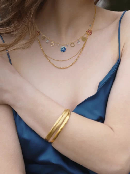 A individual in a silky blue sleeveless dress adorns layered Coral Reef Necklaces by Holly Yashi, featuring gold-plated chains with small charms. A gold bangle enhances their wrist, exuding oceanic elegance. The image is cropped to highlight the grace of the upper body and arms.