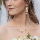 A bride in a veil and strapless dress holds pale roses, wearing Holly Yashi's Hawaiian Wedding Gold/Silver Earrings and a gold and pearl floral necklace. She smiles softly as she looks off to the side.