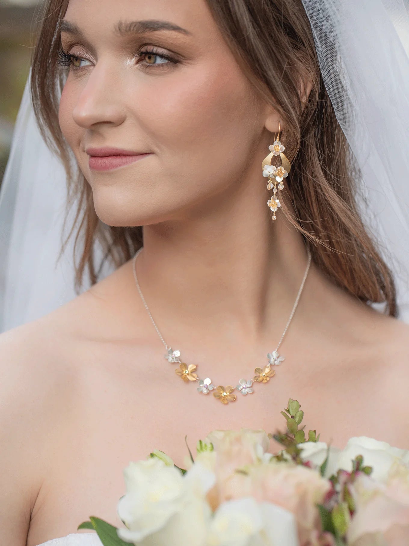A bride in a veil and strapless dress holds pale roses, wearing Holly Yashi's Hawaiian Wedding Gold/Silver Earrings and a gold and pearl floral necklace. She smiles softly as she looks off to the side.