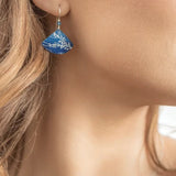A close-up of a person wearing Holly Yashis Sea Meadow Blue and Silver Earrings highlights the intricate niobium design. Their wavy, light brown hair cascades over their shoulder, exuding aquatic elegance against an out-of-focus background.