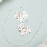 The ocean-inspired Silver Ariel Earrings by Holly Yashi, paired with two delicate silver necklaces, are beautifully showcased on a light blue surface. The hypo-allergenic shell-shaped earrings reveal a hint of white shell, evoking a serene coastal vibe.