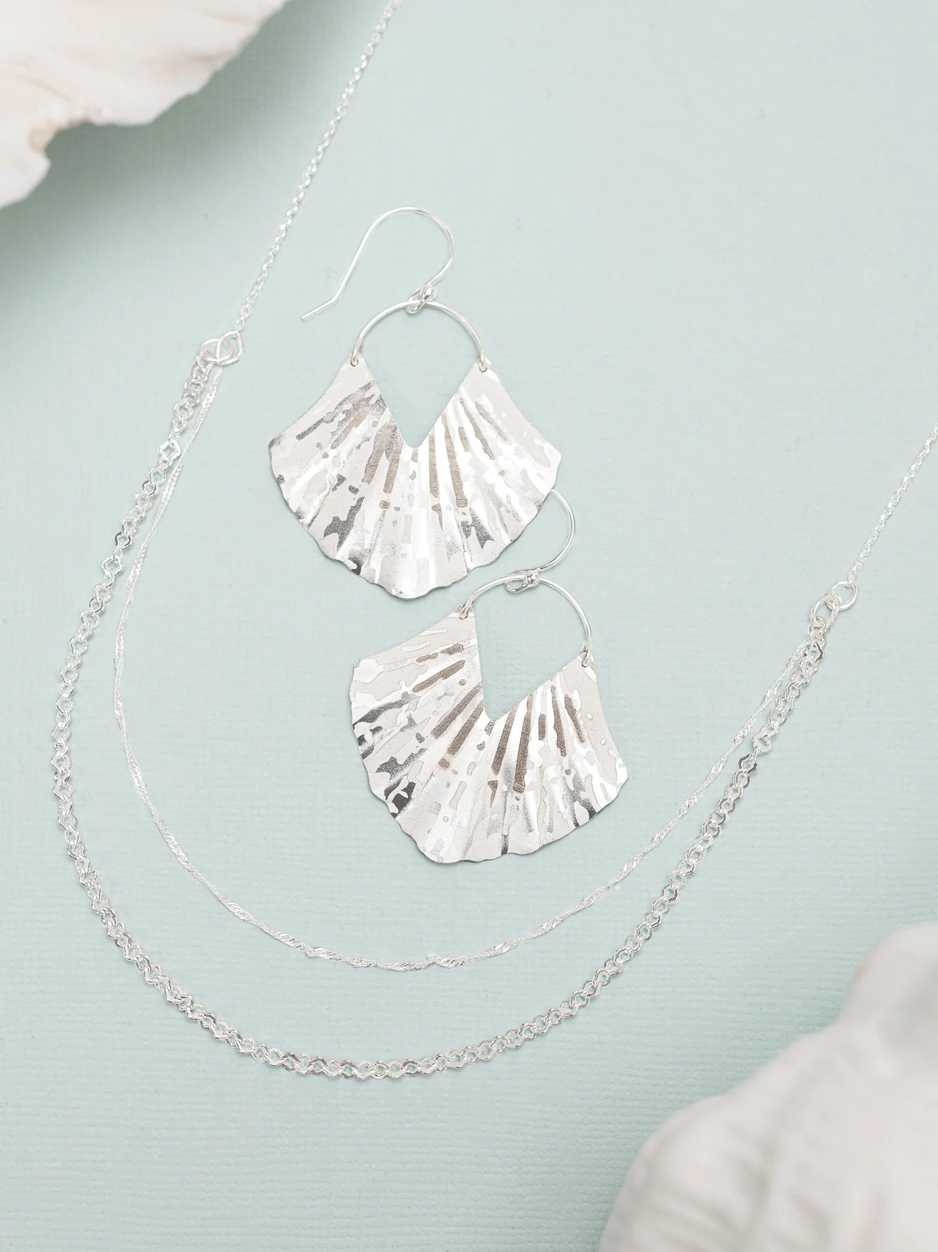 The ocean-inspired Silver Ariel Earrings by Holly Yashi, paired with two delicate silver necklaces, are beautifully showcased on a light blue surface. The hypo-allergenic shell-shaped earrings reveal a hint of white shell, evoking a serene coastal vibe.