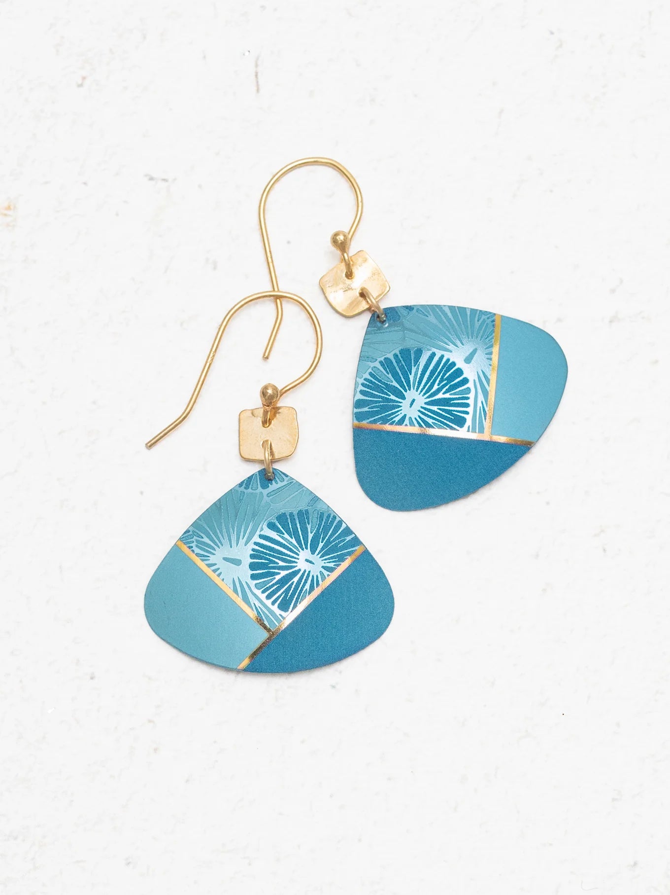 The Rowan Sky Blue Earrings by Holly Yashi feature gold hooks, geometric blue pendants, and hypoallergenic materials. Their abstract light blue, teal, and gold accents are highlighted with delicate white floral designs.