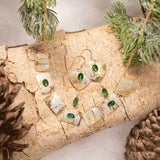 The Estelle Emerald Earrings by Holly Yashi and matching necklace are showcased on a light wood log with pine branches and pinecones, set against a snowy backdrop.