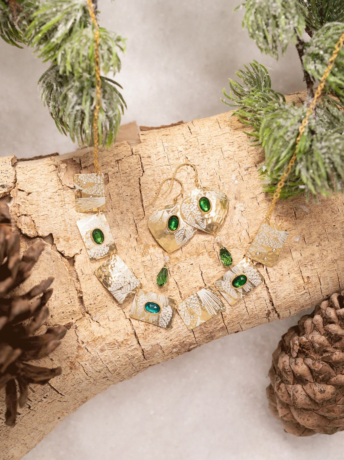 The Estelle Emerald Earrings by Holly Yashi and matching necklace are showcased on a light wood log with pine branches and pinecones, set against a snowy backdrop.