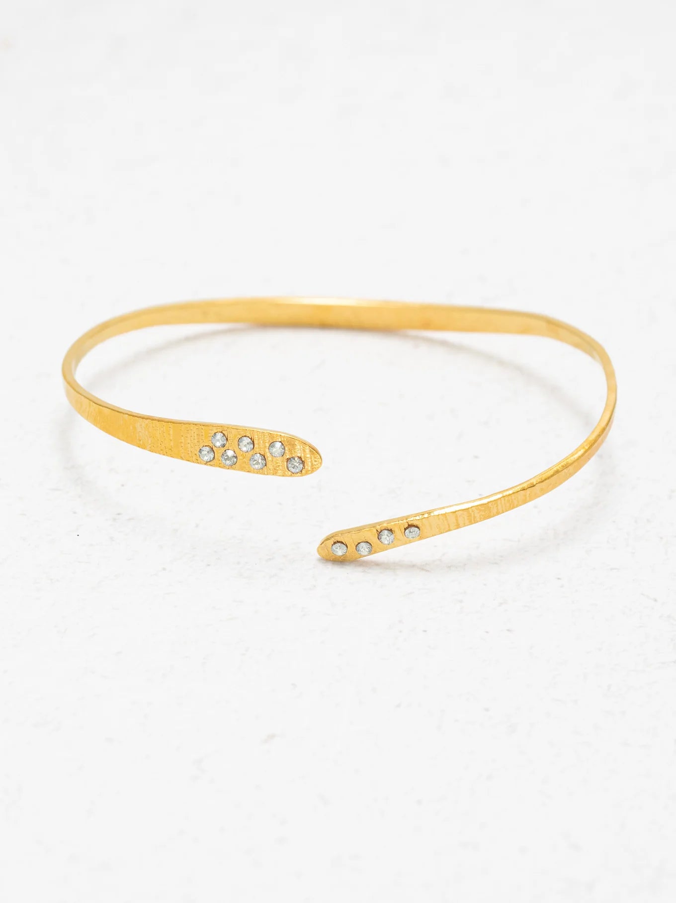 The Holly Yashi Zara Cuff Gold Bracelet is a thin, open-ended gold piece featuring small clear gemstones at each tip, shown on a white textured surface.