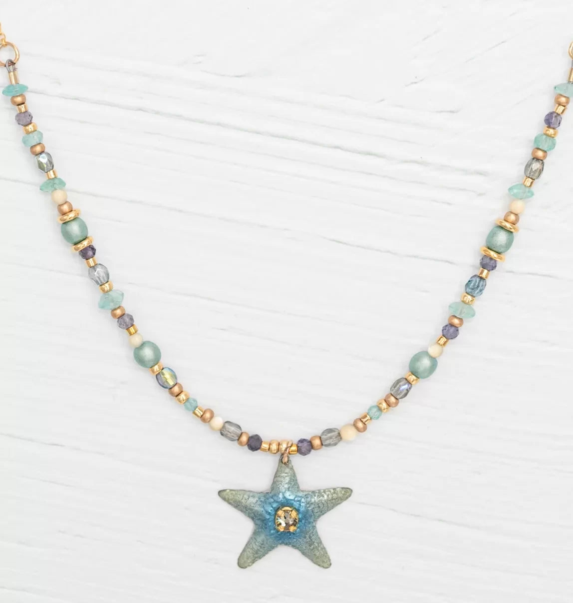 The Carmel Beaded Seashore Blue Starfish Necklace by Holly Yashi features a starfish pendant with Austrian crystals, highlighted with turquoise, gold, and blue beads. Elegantly set against a white wooden surface, it captures the serene ocean beauty.