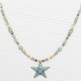 The Carmel Beaded Seashore Blue Starfish Necklace by Holly Yashi features a starfish pendant with Austrian crystals, highlighted with turquoise, gold, and blue beads. Elegantly set against a white wooden surface, it captures the serene ocean beauty.