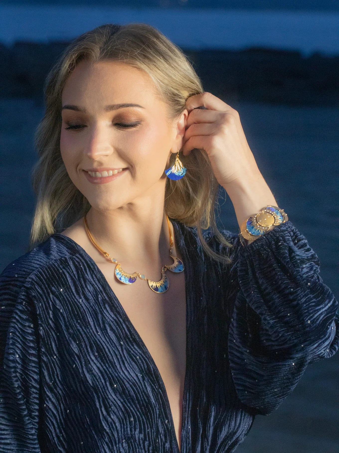 A woman in a Midnight Blue textured dress smiles with closed eyes, touching her Holly Yashi Aster Earrings in Midnight Blue. She wears a matching necklace and hypoallergenic bracelet while standing outdoors near the water at dusk.