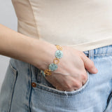 A person in light denim jeans and a beige top flaunts the Sage Oceana Bracelet by Holly Yashi, featuring 18k gold-plated textured circular charms and leaf-shaped pendants against a neutral gray background.
