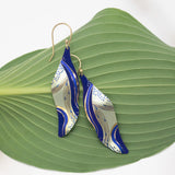 Turning Leaf Indigo Earrings by Holly Yashi, featuring abstract blue and gold leaf-shaped designs with gold-filled wires, are displayed on a green leaf against a white background.