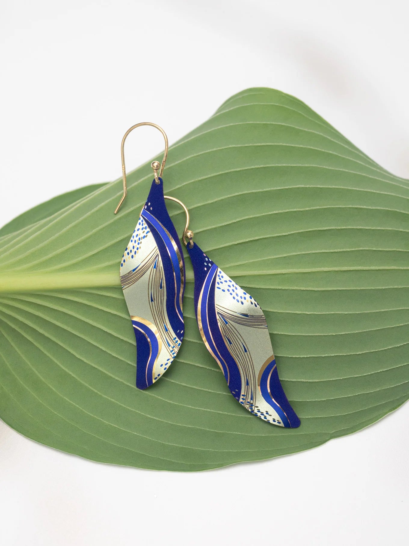 Turning Leaf Indigo Earrings by Holly Yashi, featuring abstract blue and gold leaf-shaped designs with gold-filled wires, are displayed on a green leaf against a white background.
