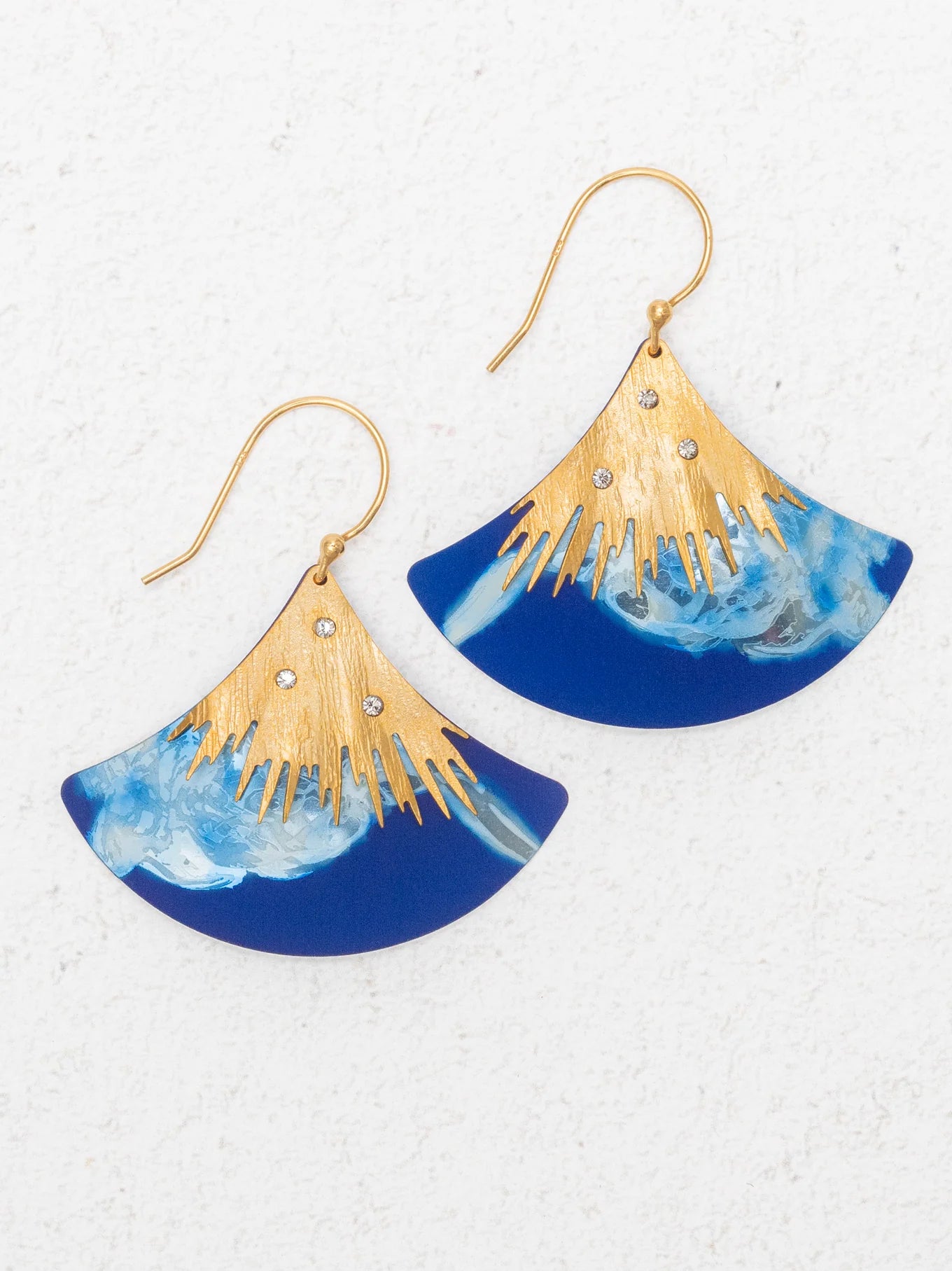The Aster Earrings/ Midnight Blue by Holly Yashi feature fan-shaped gold hooks, a gold sunburst design, rhinestones, and a marbled midnight blue lower half. Hypoallergenic for comfort. Shown on a white textured background.