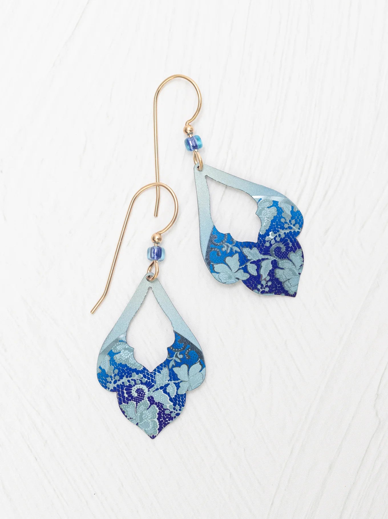 The Marisol Forever Blue Earrings by Holly Yashi feature gold hooks, hypoallergenic niobium wires, blue beads, and layered teardrop pendants with a blue floral pattern on light blue, shown on a white textured surface.