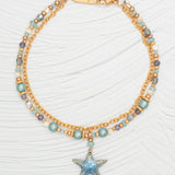 The Carmel Beaded Seashore Blue Starfish Bracelet by Holly Yashi exudes oceanic elegance with its dual gold chains adorned with blue, gold, and purple beads. A glittery blue star charm adds sparkle, while the fly engraving on the clasp completes the hypo-allergenic piece displayed on a textured white surface.