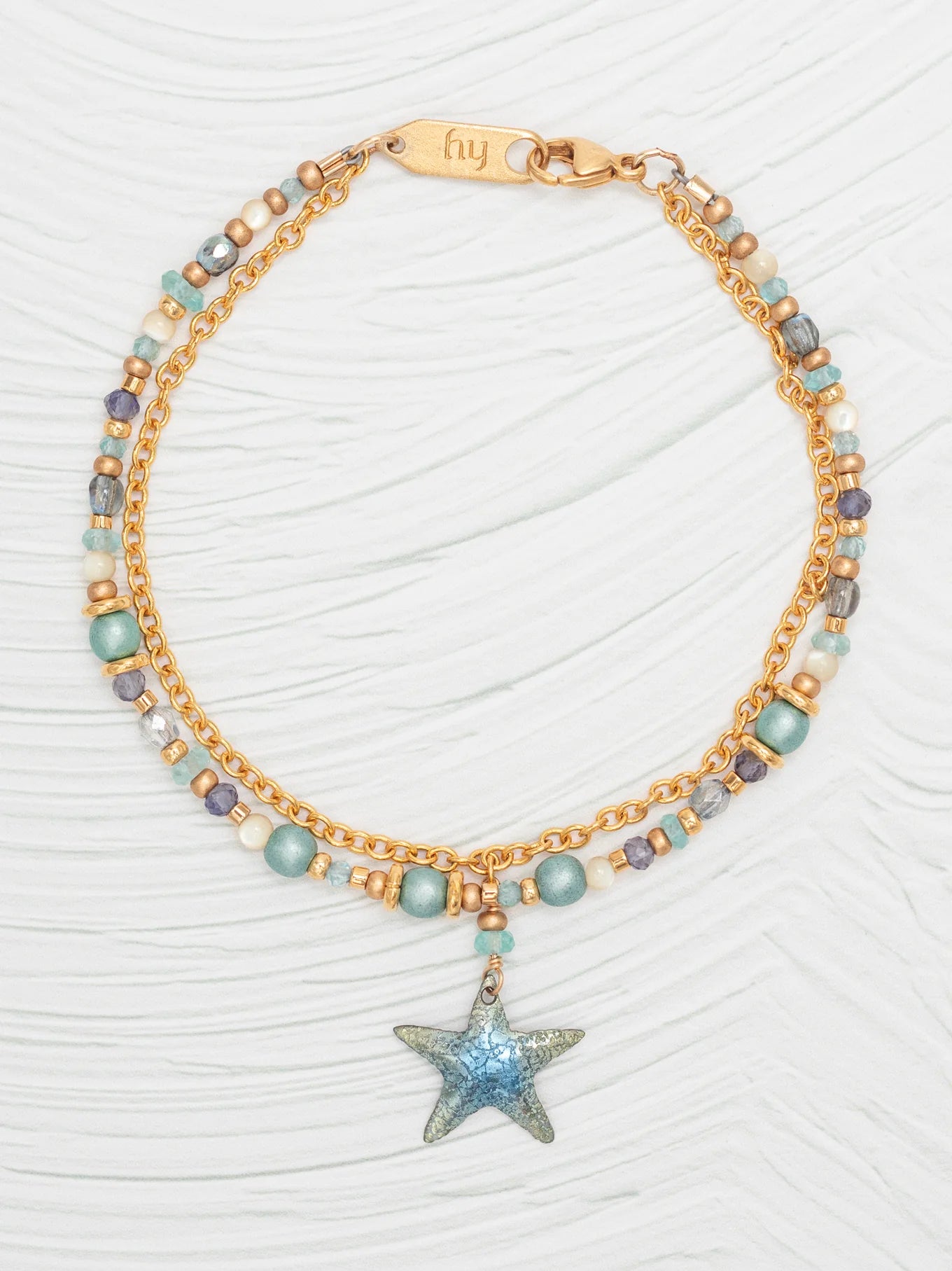 The Carmel Beaded Seashore Blue Starfish Bracelet by Holly Yashi exudes oceanic elegance with its dual gold chains adorned with blue, gold, and purple beads. A glittery blue star charm adds sparkle, while the fly engraving on the clasp completes the hypo-allergenic piece displayed on a textured white surface.