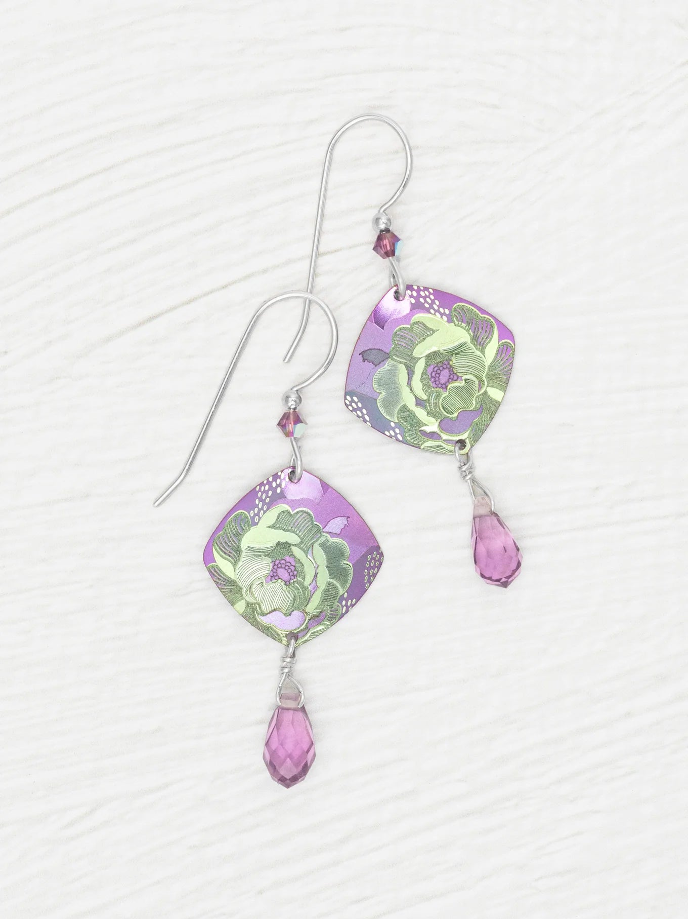 A pair of Elena Violet Earrings by Holly Yashi, featuring purple square-shaped charms with green floral designs and small purple crystal drops, displayed on a white textured surface.