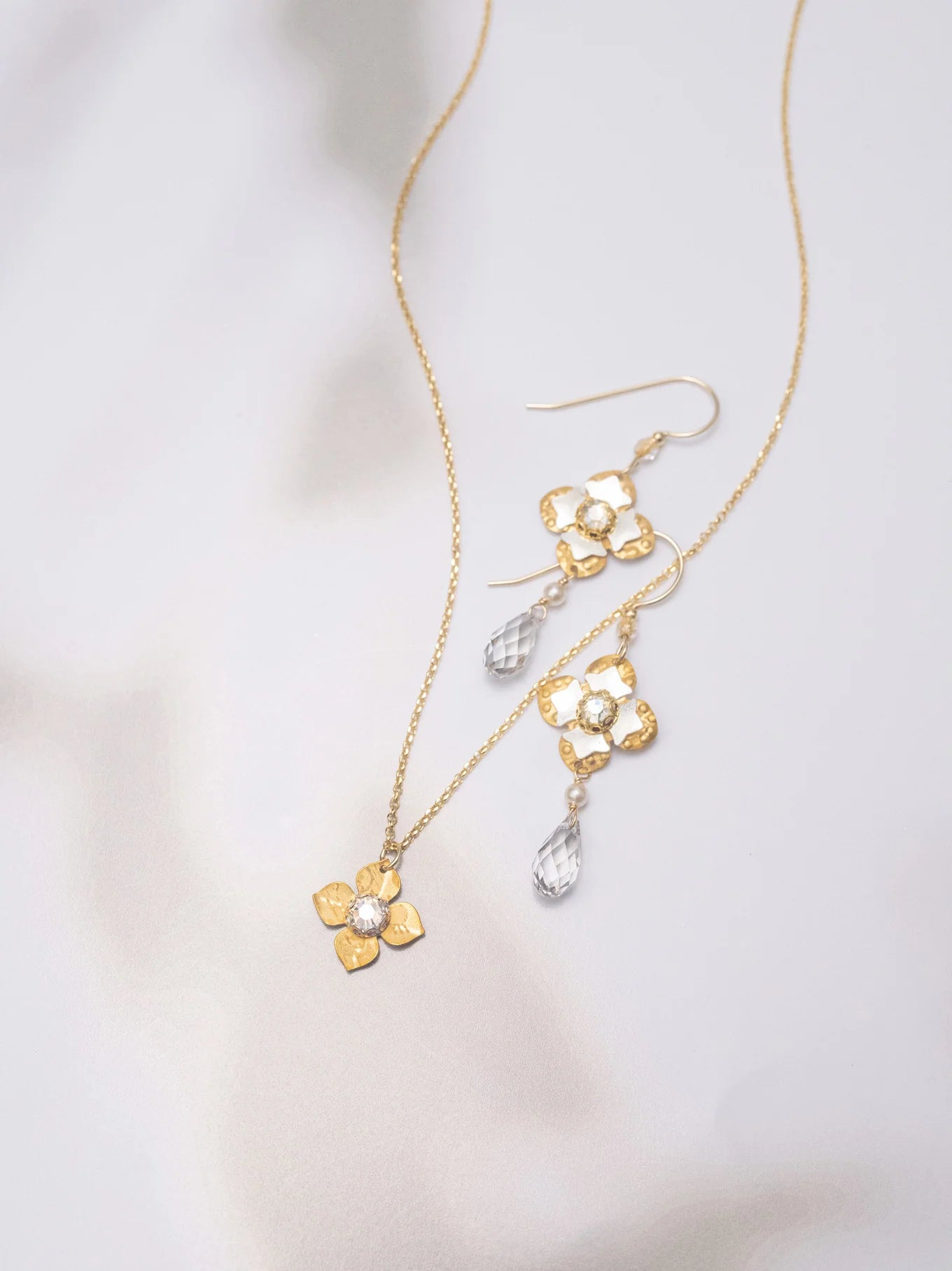 Octavia Gold Silver Earrings by Holly Yashi, inspired by Victorian style, feature clear teardrop crystals and are shown with a gold flower pendant necklace on a soft, light background.