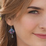 Close-up of a woman with light brown hair smiling, wearing Garden Sonnet Blue Earrings by Holly Yashi, characterized by a blue square design with vivid floral patterns and a small dangling gem against a blurred background.