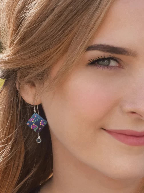 Close-up of a woman with light brown hair smiling, wearing Garden Sonnet Blue Earrings by Holly Yashi, characterized by a blue square design with vivid floral patterns and a small dangling gem against a blurred background.