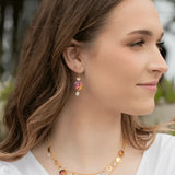 A woman with long brown hair smiles softly, wearing Coral Reef Seashore Blue Earrings by Holly Yashi and a colorful necklace. Her elegant niobium accessories complement her serene presence outdoors amid trees. She is dressed in a chic white top.