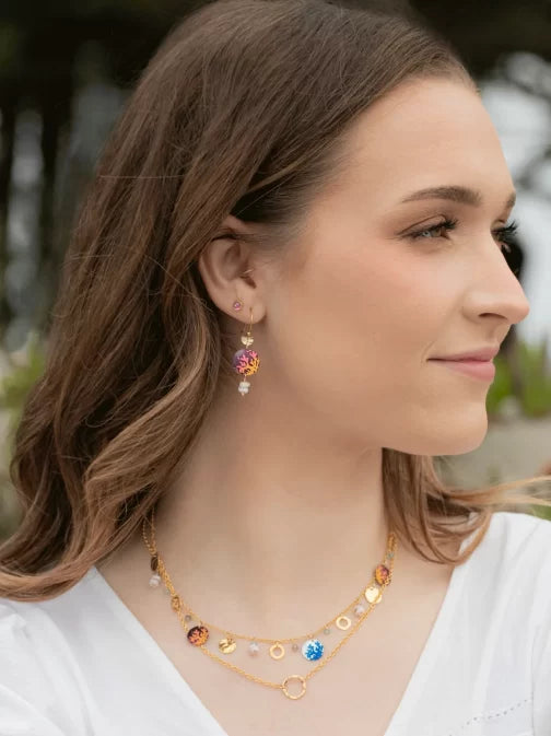 A woman with long brown hair smiles softly, wearing Coral Reef Seashore Blue Earrings by Holly Yashi and a colorful necklace. Her elegant niobium accessories complement her serene presence outdoors amid trees. She is dressed in a chic white top.