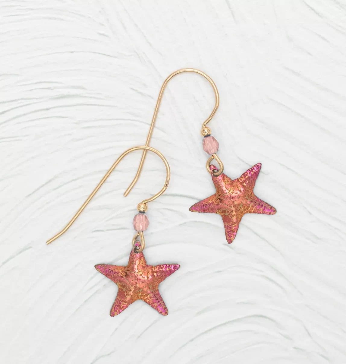Discover the Carmel Seashore Peach Starfish Earrings by Holly Yashi: gold hook earrings with pink and orange starfish charms and small pink beads. Crafted with hypo-allergenic niobium hooks, they offer an enchanting look against a textured white background.