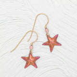 Discover the Carmel Seashore Peach Starfish Earrings by Holly Yashi: gold hook earrings with pink and orange starfish charms and small pink beads. Crafted with hypo-allergenic niobium hooks, they offer an enchanting look against a textured white background.