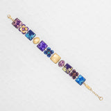 The Eliza Tiled Multi-Color Bracelet by Holly Yashi features a gold chain with square and round charms in purple, blue, and pink, each adorned with floral and abstract designs, displayed on a white textured surface.