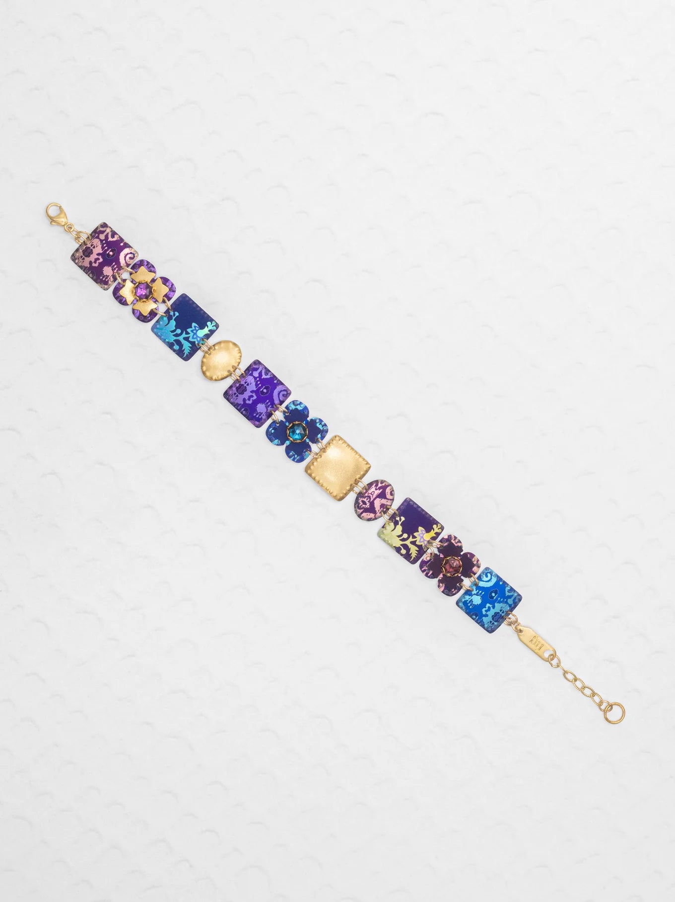 The Eliza Tiled Multi-Color Bracelet by Holly Yashi features a gold chain with square and round charms in purple, blue, and pink, each adorned with floral and abstract designs, displayed on a white textured surface.
